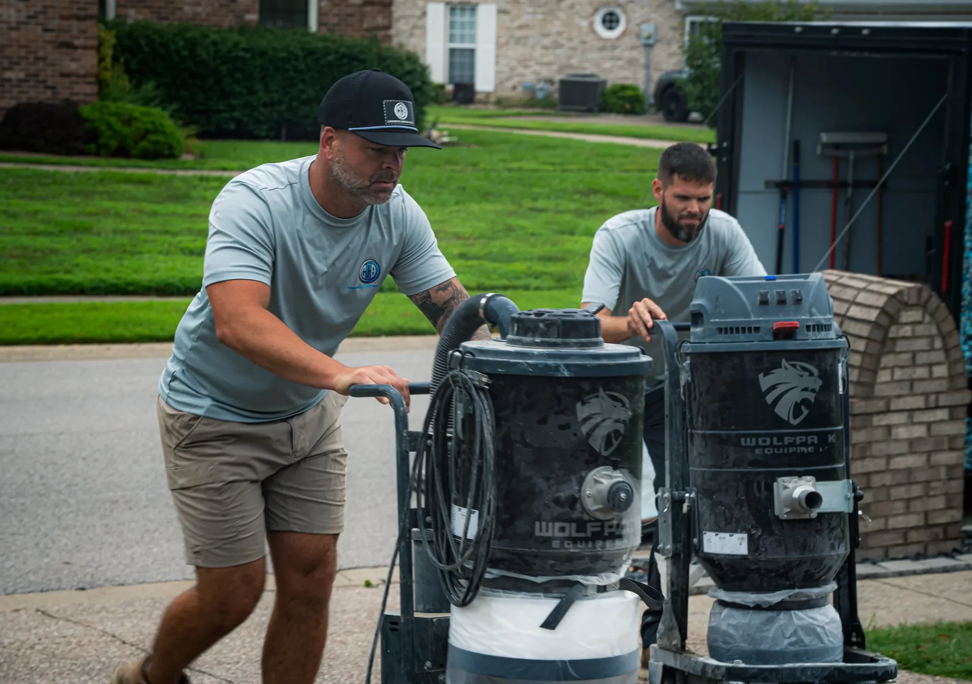Two Concrete Coating Bros team members in branded shirts wheeling industrial vacuum equipment along a residential street