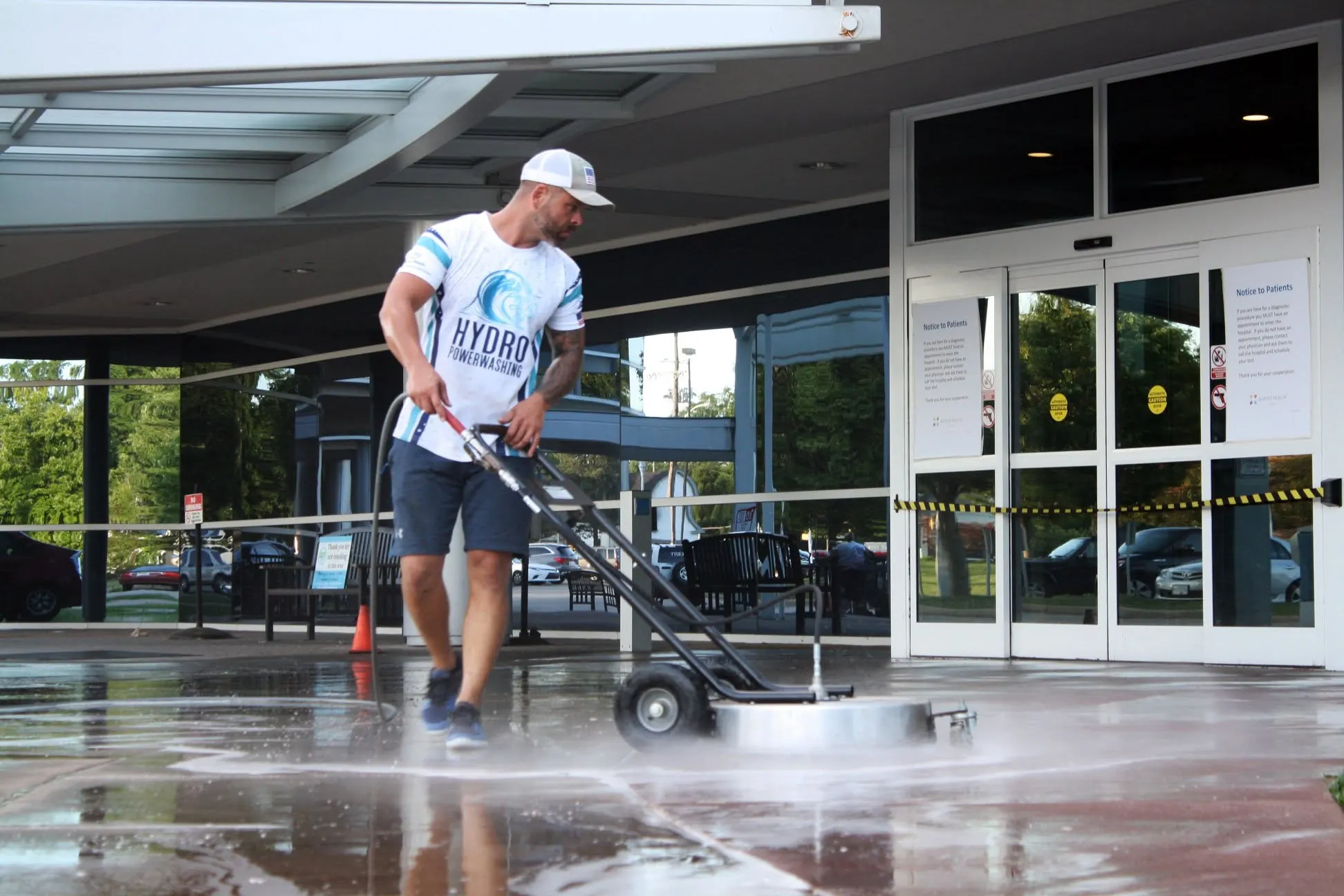 Worker pushing a rolling surface cleaner across wet concrete under a commercial entrance canopy