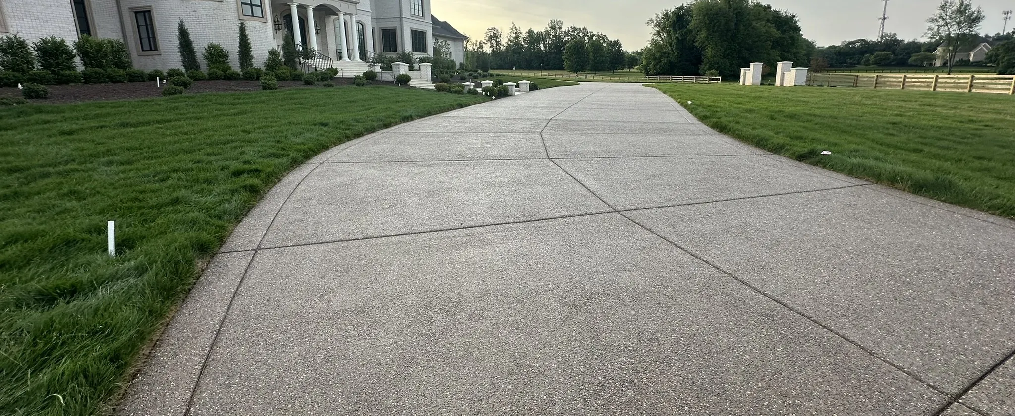 Long exposed-aggregate concrete driveway with saw-cut joints curving between lawns toward a white brick home