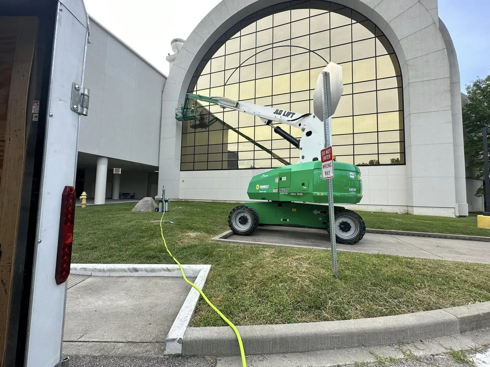 Green boom lift on a lawn in front of a building with a large arched window
